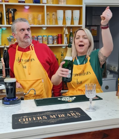 Man and woman behind the Cheers m'dears bar, raising a drink in celebration