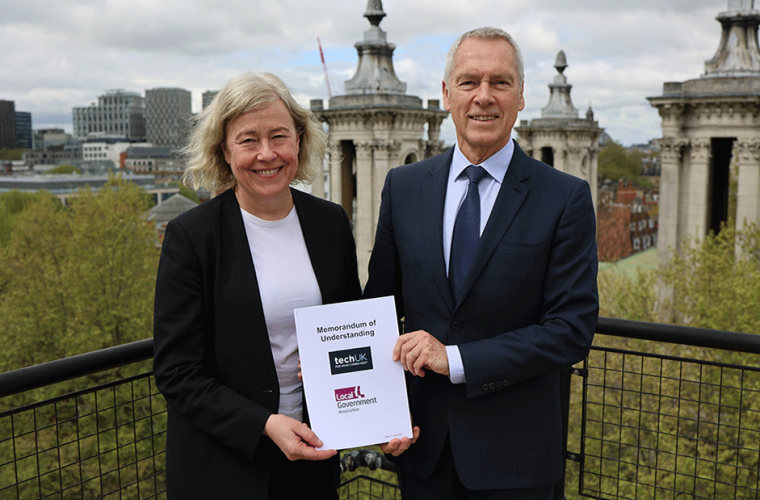 LGA Chief Executive Joanna Killian with techUK CEO Julian David stand holding the memorandum of understanding between them with the London skyline behind them and smiling at the camera
