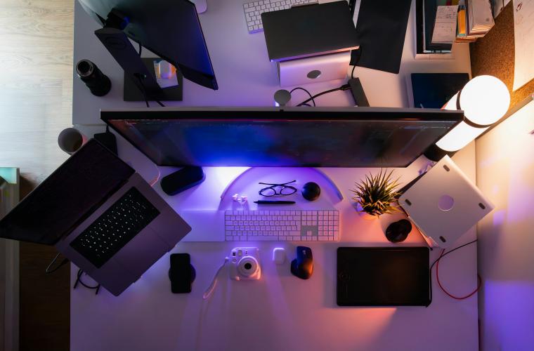 A modern home office desk setup with multiple monitors, a laptop, keyboard, camera, notebooks, and other tech accessories, illuminated by colorful blue and orange lighting.